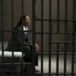Young businesswoman sitting on bed in prison cell, looking away