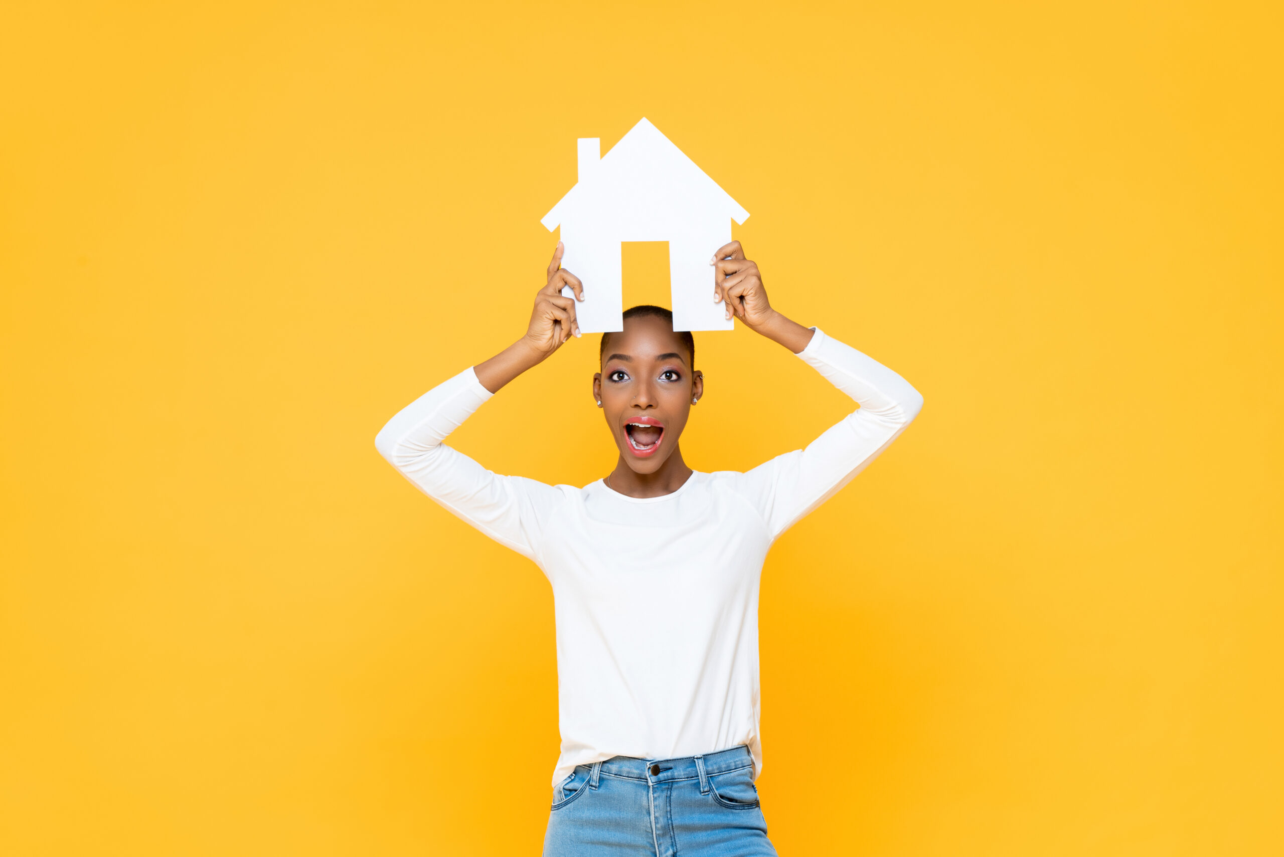 Surprised African American woman holding house sign overhead isolated on yellow background